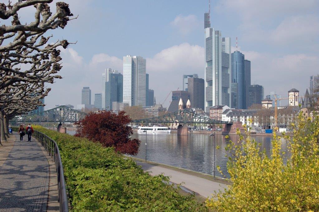 Das Bild zeigt die Skyline von Frankfurt am Main mit dem Main und Bäumen im Vordergrund sowie den Hochhäusern dahinter.