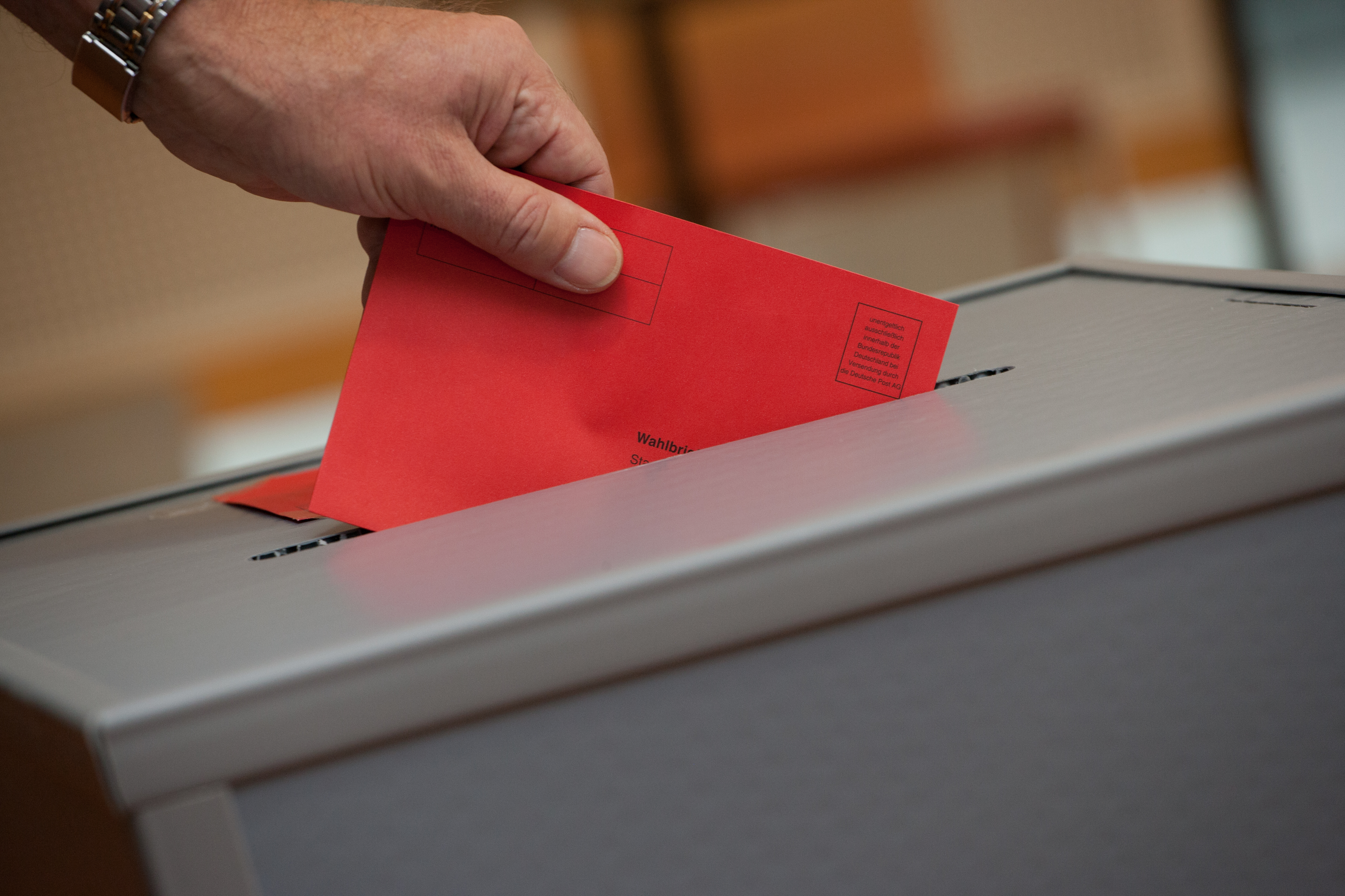 Eine Wahlurne, in der der Stimmzettel eingeworfen wird. Die Wahlurne ist grau, der Stimmwahlumschlag ist rot.