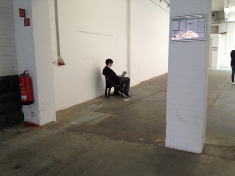 Das Bild zeigt eine leere Werkhalle mit weißen Wänden, hier sitzt einsam ein Mensch mit einem Laptop auf den Knien auf einem Hocker sitzend, mit dem Rücken zum Fotografen.