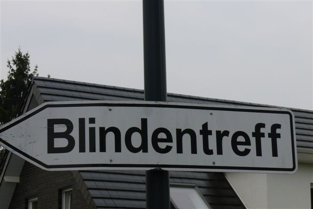 Das Bild zeigt ein Verkehrsschild mit der Aufschrift "Blindentreff", es hängt an einem Verkehrsmast gut sichtbar, im Hintergrund ist ein Häuserdach zu erkennen sowie grauer Himmel darüber.