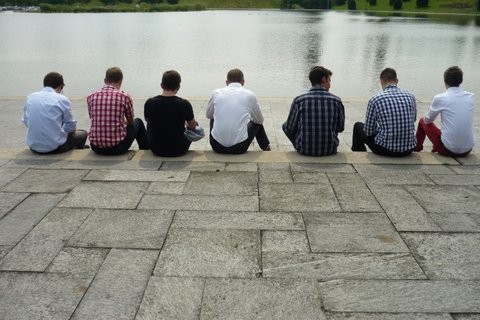 Das Foto zeigt junge Männer, die in einer Reihe an einem See sitzen, sie sind aber von hinten fotografiert.