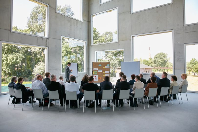 Zwei Dutzen Workshop-Teilnehmer sitzen in einem Halbkreis vor einer großen Glasfront. Vorne zu sehen ist ein Moderator mit einer Metplanwand und einer Flipchart