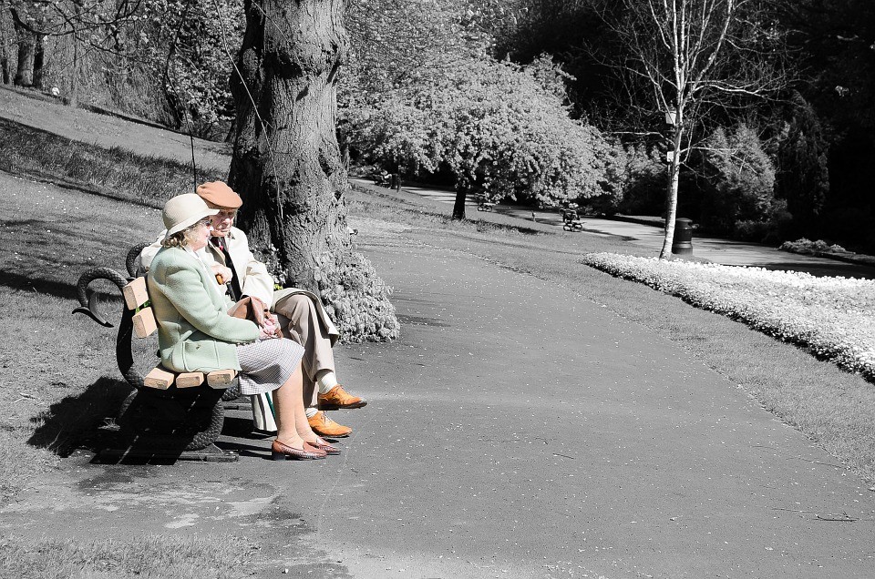 Ein älteres Ehepaar sitzt in einem Park auf einer Bank. Ihre Kleidung ist farbig, alles um sie herum schwarz und weiß