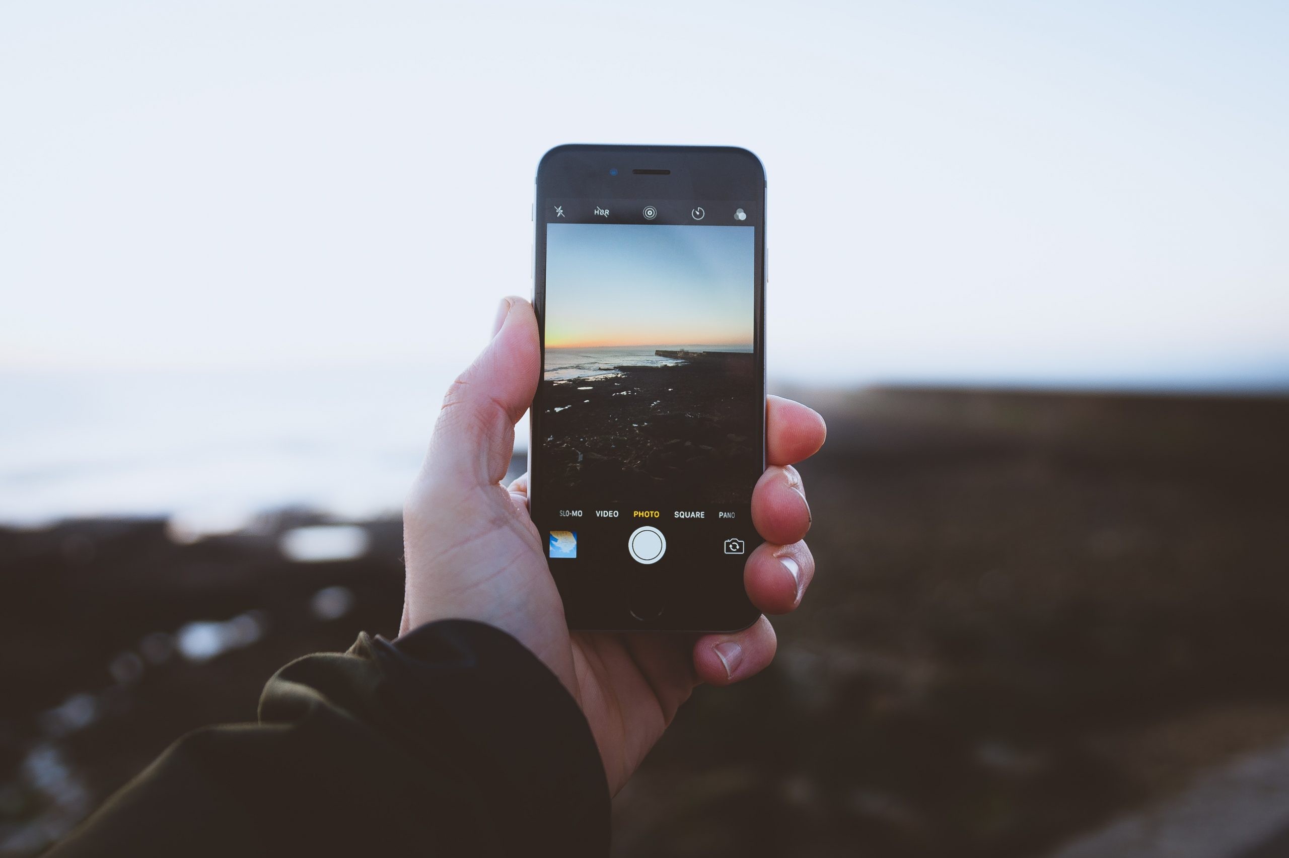 Eine Hand hält ein Smartphone auf das Meer. Zu sehen ist durch das Display das Meer und die Küste