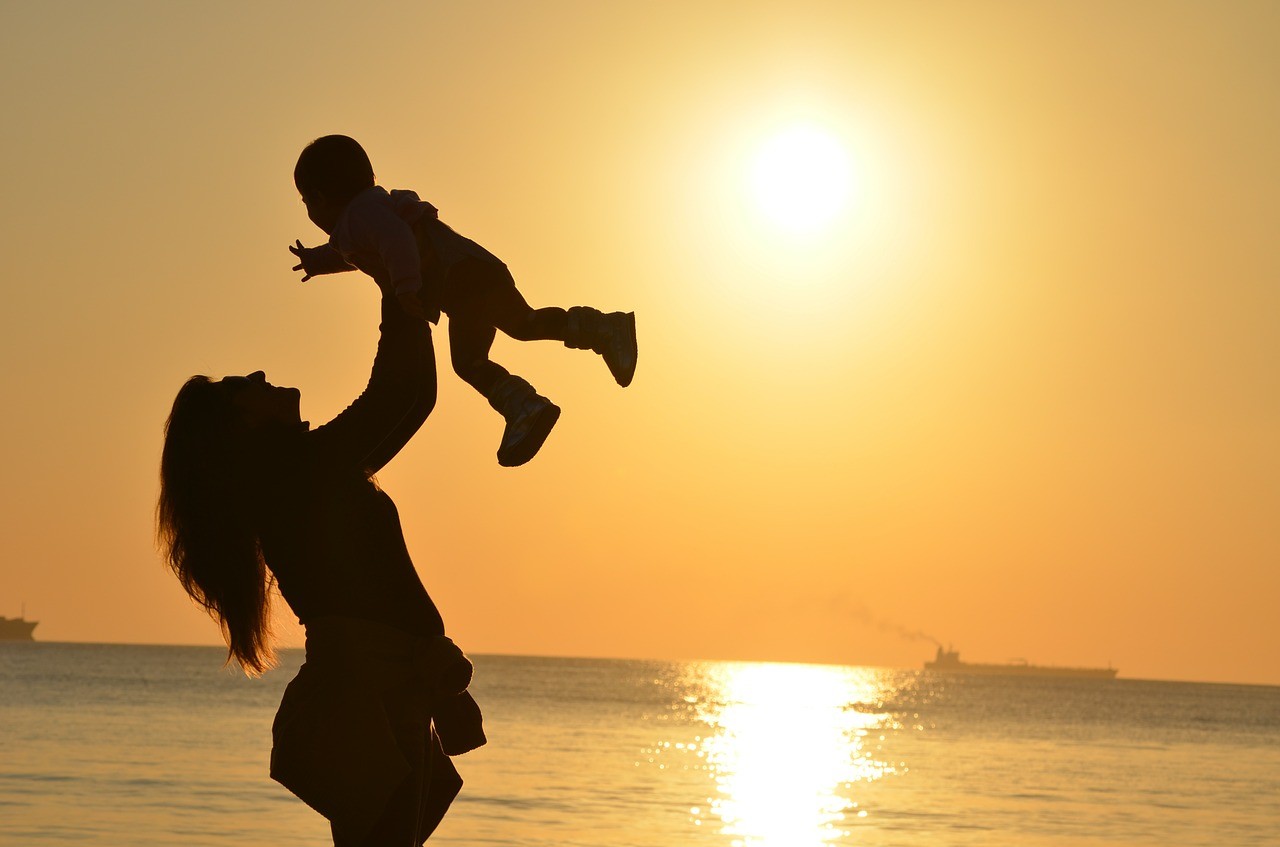Eine Frau hält am Meer ein Baby in die Höhe. Die Sonne geht unter und die Frau und das Baby sind durch die glühende Sonne im Hintergrund nur in schwarz zu sehen.