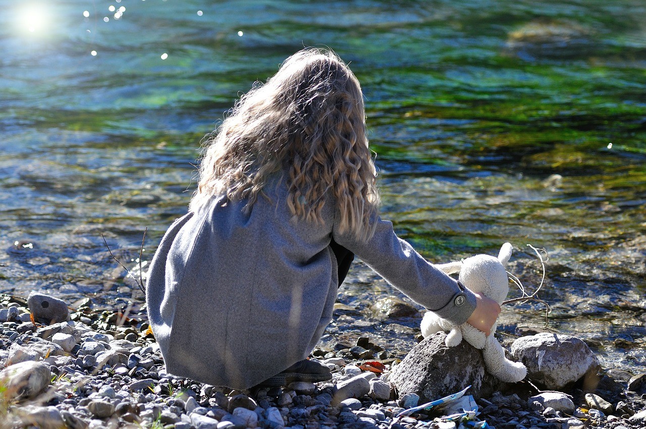 Ein Kind hockt mit einem Kuscheltier am Ufer eines Flusses. Man sieht nur den Rücken des Mädchens.