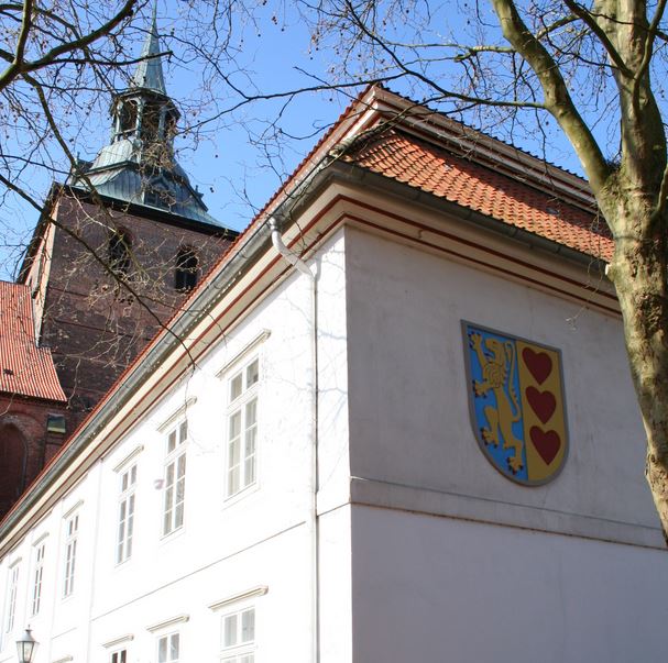 Das Gebäude der Landkreisverwaltung Lüneburg. Im Vordergrund ein weißes Gebäude mit dem Kreiswappen, im Hintergrund ein Kirchturm