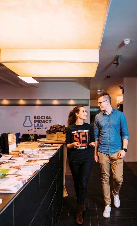 Ein junger Mann und eine junge Frau unterhalten sich im Gehen in einem großen Büroraum. Im Hintergrund das Schild: Social Impact Lab