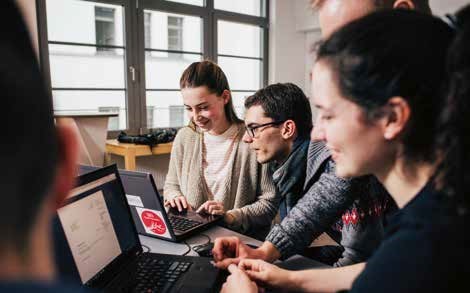 Mehrere junge Leute stehen um zwei Laptops herum. Alle sind am Lächeln.